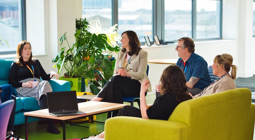 A group of PSS teammates are sat together on bright coloured sofas. They are all smiling.