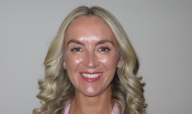 The picture is of Sarah on a white background. She has blonde hair to her shoulders and is wearing a pink top. She is smiling.