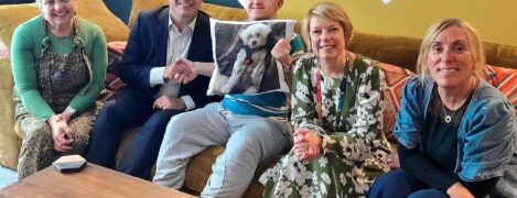 Shared Lives is an alternative form of adult social care. PSS Shared Lives carer, Sally Smith, MP Aaron Bell, supported person Paul with PSS UK CEO, Lesley Dixon and Registered Manager Lisa Hill. They sit on an extra large couch together and Paul is holding up a pillow with a picture of their dog on it.
