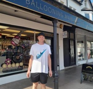 Paul stands in front of Ballooning Marvellous, the shop owned by his Shared Lives carer, Andrea. It's sunny and he's in a t shirt, shorts, and cap