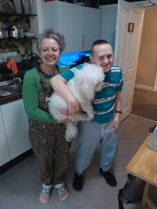 Paul who has downs syndrome poses with his shared lives carer sally, who is also holding their white fluffy dog, Haatchi - they are in the kitchen and smiling 