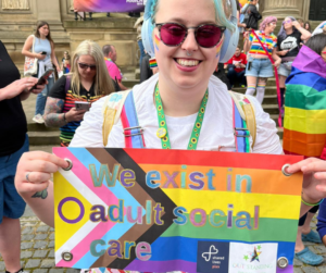 Hope has blue hair and purple tinted glasses on - she smiles proudly as she holds a sign that says 'we exist in adult social care' and she's wearing a white t shirt and a rainbow coloured dungarees