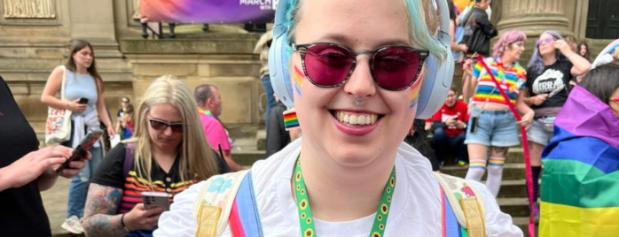 Hope has blue hair and purple tinted glasses on - she smiles proudly as she holds a sign that says 'we exist in adult social care' and she's wearing a white t shirt and a rainbow coloured dungarees