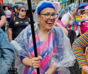 Mandy smiles at the camera - she's pictured at Pride holding the pole to the banner and she's wearing a dress with the bisexual flag colours. She wears black rimmed glasses and a blue headband to match the blue on her dress.