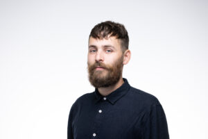 Tom is a slim white male with a big beard and brown hair - he is wearing a navy buttoned up shirt and the photo is quite professional - like a headshot. Tom looks really calm and approachable.