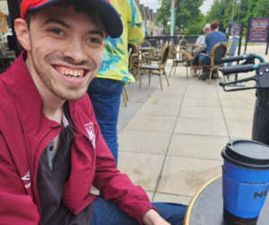 James is out and about in his local high street sat down drinking a coffee. he's outdoors al fresco style - he's smiling widely at the camera and wearing a cap and a read sports jacket.