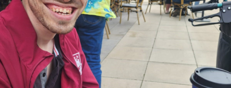 James is out and about in his local high street sat down drinking a coffee. he's outdoors al fresco style - he's smiling widely at the camera and wearing a cap and a read sports jacket.